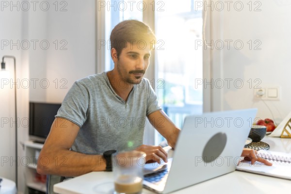 Young man working from home, typing on a laptop and jotting down notes while basking in the warm morning sunlight streaming through the window