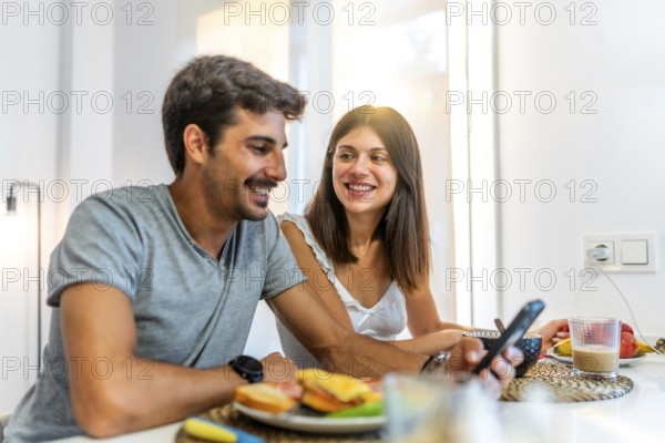 Young couple smiling and enjoying breakfast together in their cozy kitchen, using a smartphone to connect and share moments online