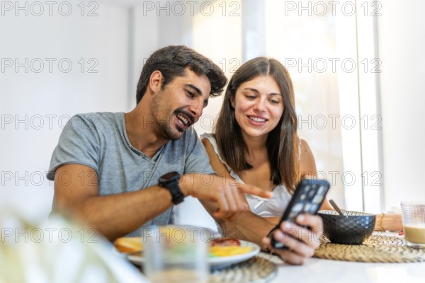 Young couple enjoying breakfast at home, smiling and pointing at a smartphone while browsing social media together at the kitchen table