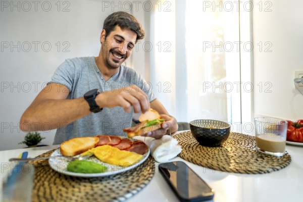 Young man smiling while preparing a delicious sandwich for breakfast at home, enjoying a healthy start to the day in a bright kitchen