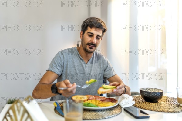 Man adding fresh avocado to toast while enjoying a healthy breakfast at home, embracing a balanced morning routine in a modern kitchen