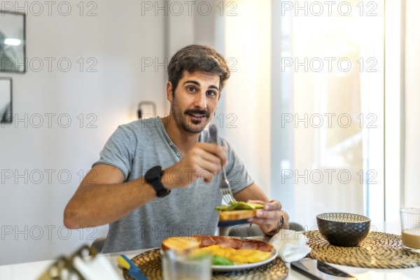 Happy man enjoying avocado toast alongside ham and eggs while relaxing in his modern apartment, soaking in the morning light