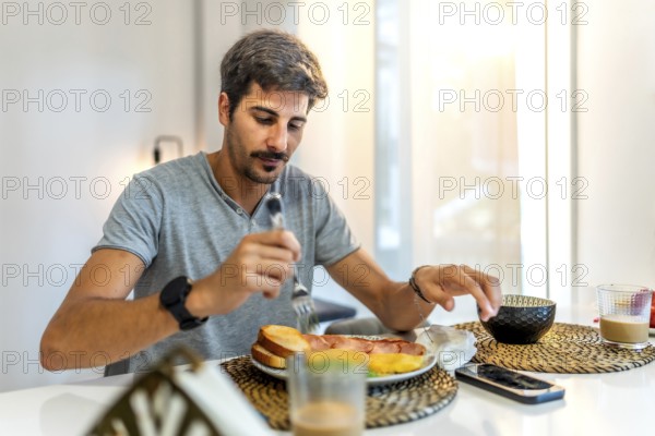 Young man enjoying breakfast with crispy bacon, fluffy eggs, toast, and steaming coffee in a stylish modern kitchen setting
