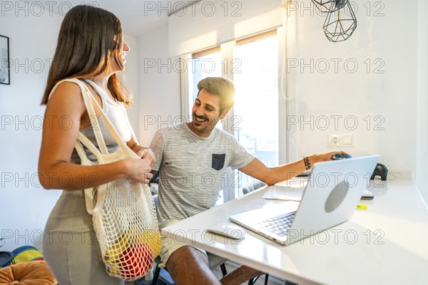 Young woman arriving home from grocery shopping, showing fruits and vegetables in reusable bag to her boyfriend working from home
