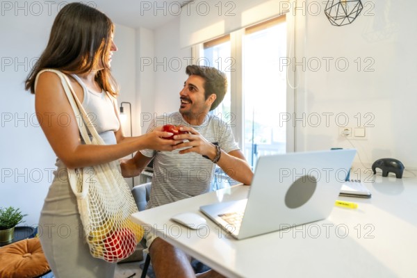 Young woman arriving home from grocery shopping, giving an apple to her boyfriend working remotely