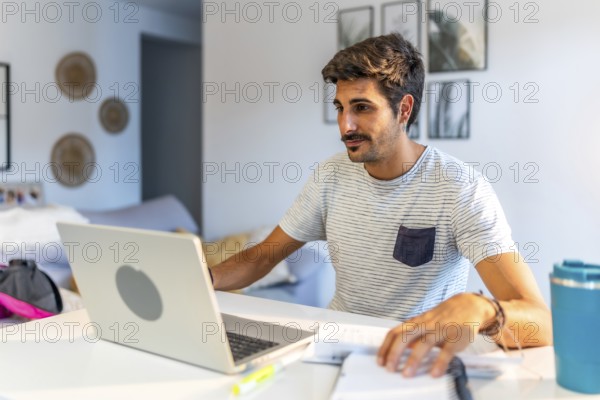 Young male freelancer working remotely at home, using a laptop and reviewing notes while sitting comfortably at his desk