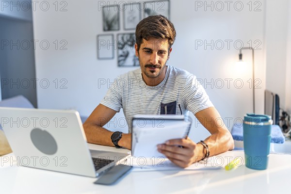 Young man managing his business remotely, reviewing notes and working on his laptop in his bright home office