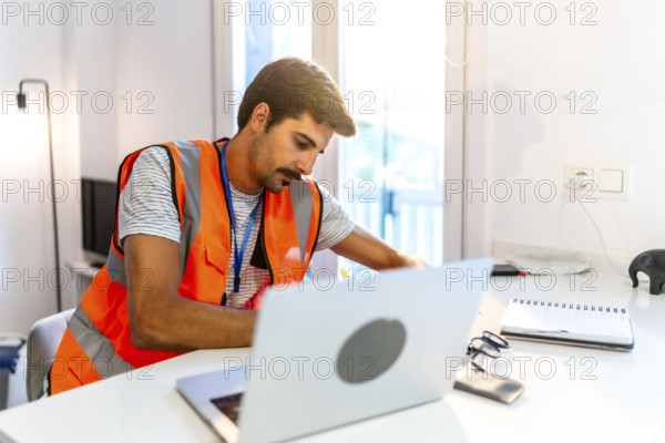 Focused engineer wearing a high visibility vest working from home, using a laptop and taking notes in a bright, modern home office