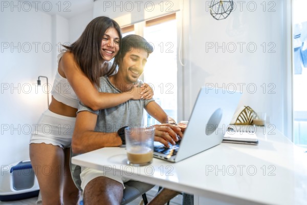 Young smiling couple using laptop at home, woman hugging man working on computer, modern bright apartment, love and technology