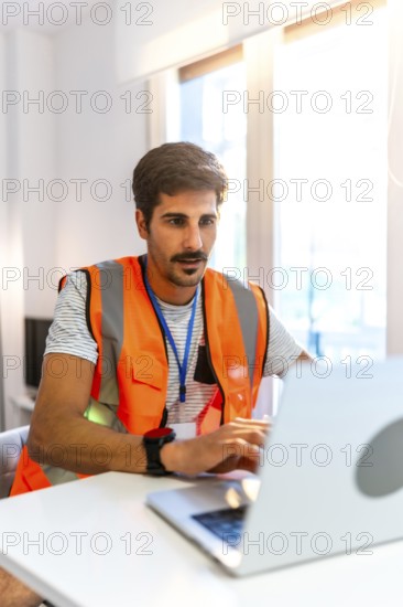 Focused engineer wearing high visibility vest working on laptop in bright modern office, managing projects and ensuring workplace safety