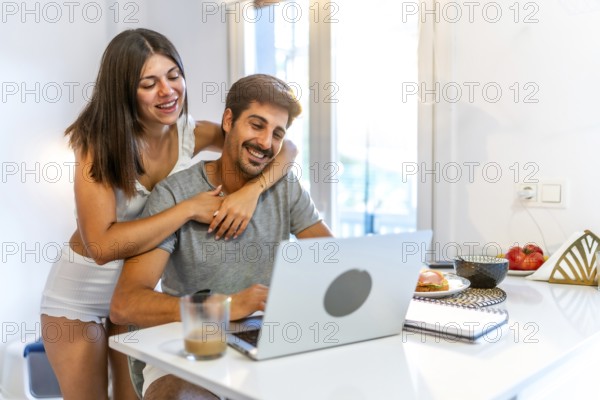Young couple embracing and enjoying each other's company while working on a laptop in a cozy kitchen filled with natural light