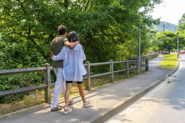 Romantic couple walking on a paved path in a park, embracing each other, enjoying a sunny summer day