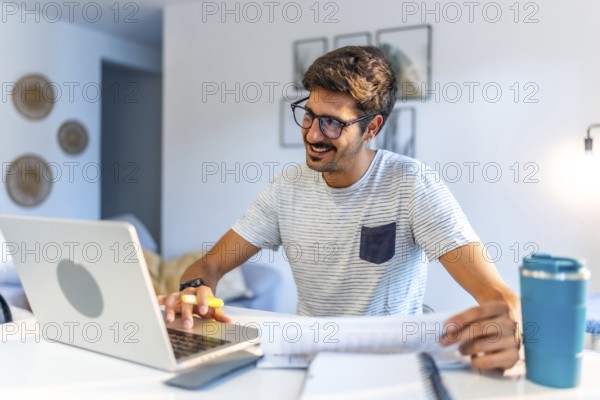 Young man working comfortably at home, engaging with a laptop and reviewing documents, smiling while enjoying a productive day