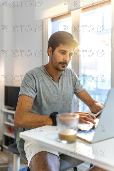 Young man concentrating on his laptop, working remotely from his bright home office while enjoying a cup of coffee