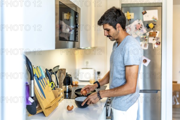 Young man cracking eggs into a bowl, preparing to cook breakfast or a meal in his modern, well equipped kitchen