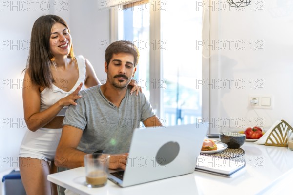 Young woman massaging her husband's shoulders while he's working from home on his laptop in the kitchen