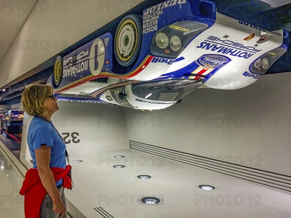 Visitor to the museum looks upwards at a Porsche 956 racing car hanging upside down under the ceiling of the museum as an example of high contact pressure at 321.4 kilometres per hour, Porsche Museum, Stuttgart, Baden-Württemberg, Germany