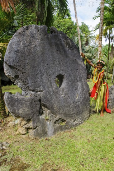 Local boy in traditional traditional costume from Yap Island as size comparison next to oversized large huge coins made of stone real means of payment in stone money bank stone money bank in jungle of Yap Island, Yap Island, Yap State, Caroline Islands, FSM, Federated States of Micronesia, Australia, Oceania