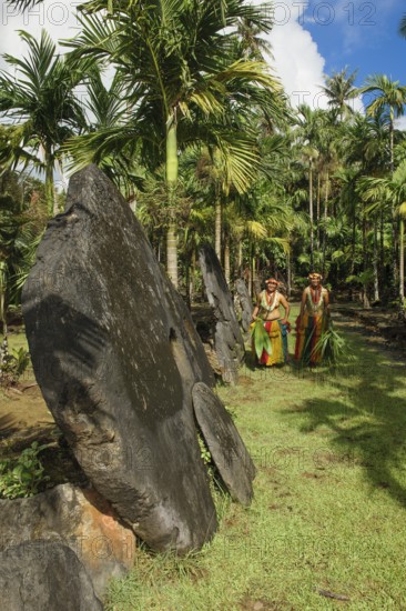 Native couple from Yap in traditional traditional costume walking on path through palm forest passing oversized large huge stone coins real currency in stone money bank stone money bank in jungle of Yap Island, Yap Island, Yap State, Caroline Islands, FSM, Federated States of Micronesia, Australia, Oceania