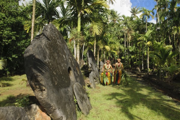 Native couple from Yap in traditional traditional costume walking on path through palm forest passing oversized large huge stone coins real currency in stone money bank stone money bank in jungle of Yap Island, Yap Island, Yap State, Caroline Islands, FSM, Federated States of Micronesia, Australia, Oceania