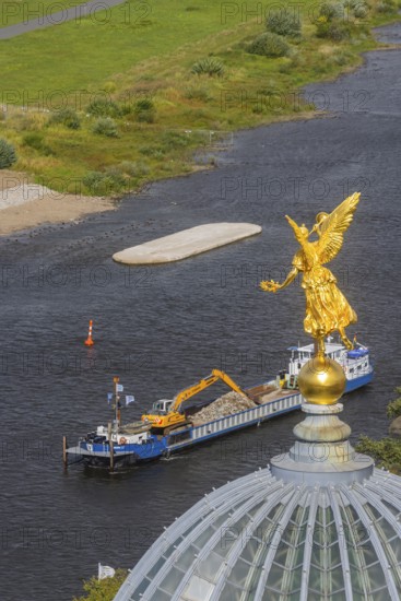 View from the lantern of the Church of Our Lady. Fama (the goddess of rumours) on the dome of the Academy of Fine Arts, with the pillar foundations of the former Carola bridges. The dredger Domarin straightens the fairway for shipping, Dresden, Saxony, Germany