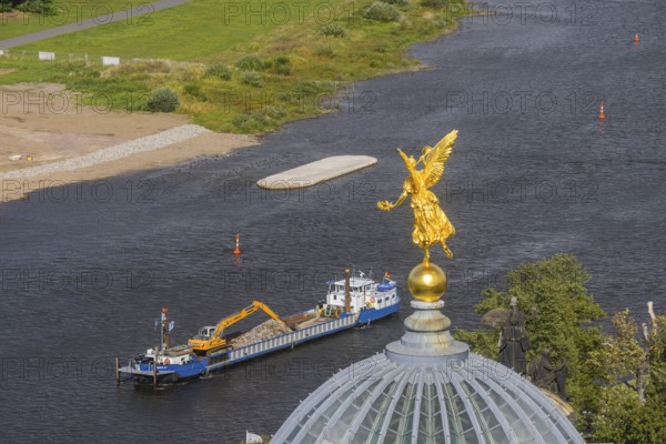View from the lantern of the Church of Our Lady. Fama (the goddess of rumours) on the dome of the Academy of Fine Arts, with the pillar foundations of the former Carola bridges. The dredger Domarin straightens the fairway for shipping, Dresden, Saxony, Germany