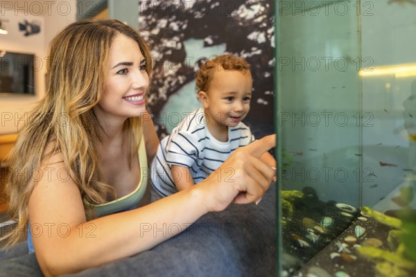 Happy mother and her baby son pointing at colorful fish swimming in a home aquarium, enjoying quality time together