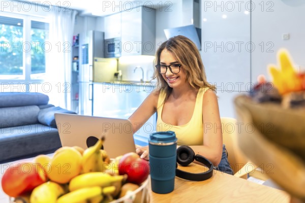 Young woman working remotely from her home office using laptop, enjoying healthy lifestyle with fresh fruit on the table