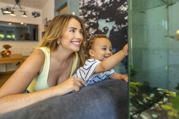 Happy mother and her baby son lying on the sofa, enjoying a delightful moment together while watching the colorful fish in the aquarium