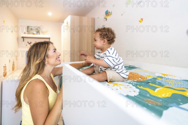 Smiling mother looking at her cute baby sitting on a bunk bed in a decorated bedroom, happy family concept