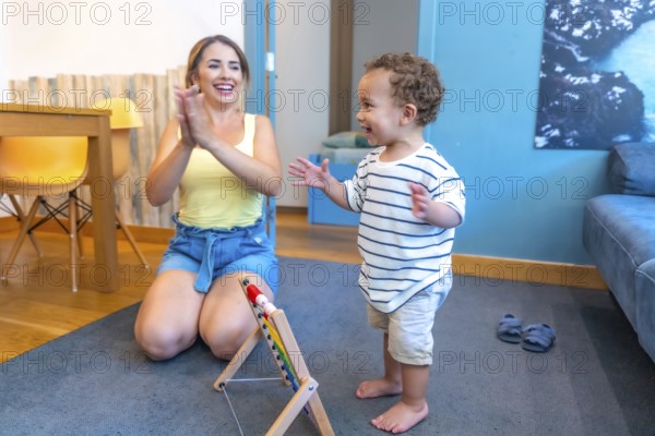 Mother encouraging her baby boy to walk, clapping hands in living room, promoting child development and early childhood education