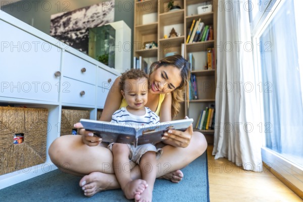 Happy mother spending quality time reading a book to her baby son, sitting on the floor in the living room
