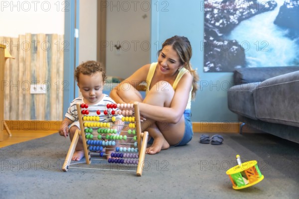 Happy mother watching her baby boy playing with a colorful abacus, developing early math skills in a playful home environment