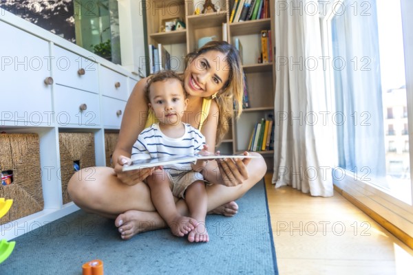 Happy mother and her baby son reading a book together, sitting on the floor in the living room at home