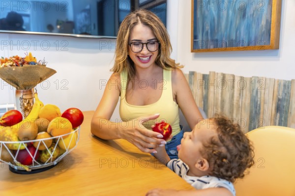 Loving mother giving a fresh peach to her baby son while sitting at the kitchen table, promoting healthy eating habits