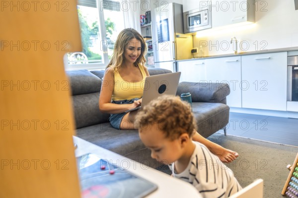 Woman telecommuting on her laptop while her baby plays nearby in a modern living room, blending work and family life seamlessly