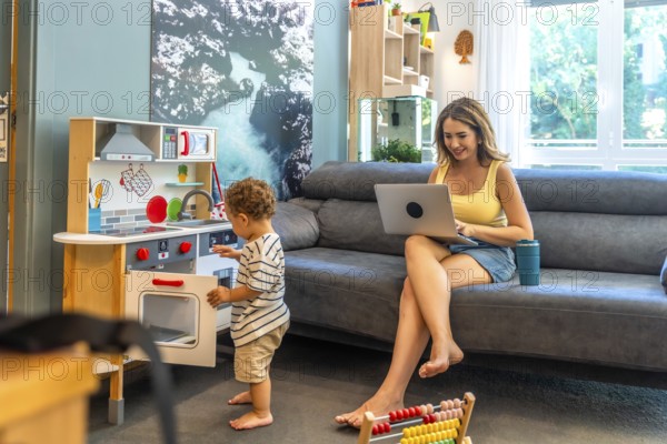 Woman working remotely on laptop while her toddler plays with a toy kitchen, demonstrating work life balance in a home setting