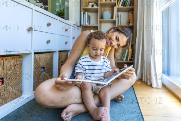 Young mother sitting on the floor reading a book with her little son, enjoying quality time together in their cozy living room