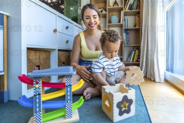 Smiling mother watching her cute toddler son playing with a shape sorter toy and a colorful car ramp toy in a cozy playroom