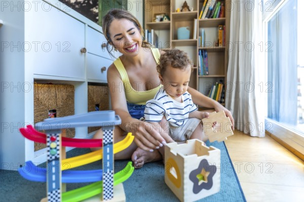 Happy mother and son are sitting on the floor playing with a wooden shape sorter and colorful toy car ramp in a cozy kids room