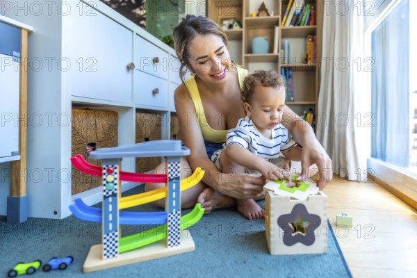 Happy mother helping her baby son playing with a shape sorter toy, sitting on a play mat in a modern living room