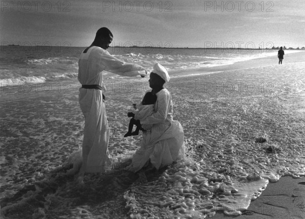 NGA, Lagos, 20.05.97 -- Free churches in Lagos. -- A priest uses the water of the Atlantic Ocean to baptize a child who is being held by his mother. During their religious practice, believers wear white robes, the clothes with which they want to stand in front of God.