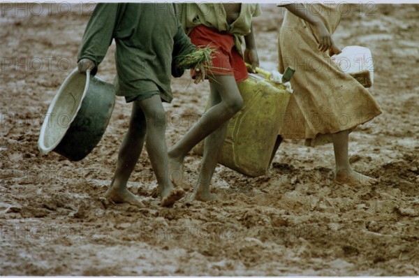 RWA, Benaco camp, 19.05.1994 -- Refugee children wander through the muddy paths of the Benaco camp to get food and water. -- Rwandan refuggee children walk a muddy road for their daily food and water supply at the Benaco camp.