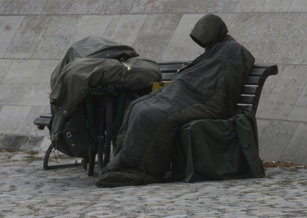 DEU Germany Germany Berlin A homeless person sits on the banks of the Spree opposite the Federal Chancellery, wrapped in his sleeping bag, his bicycle covered