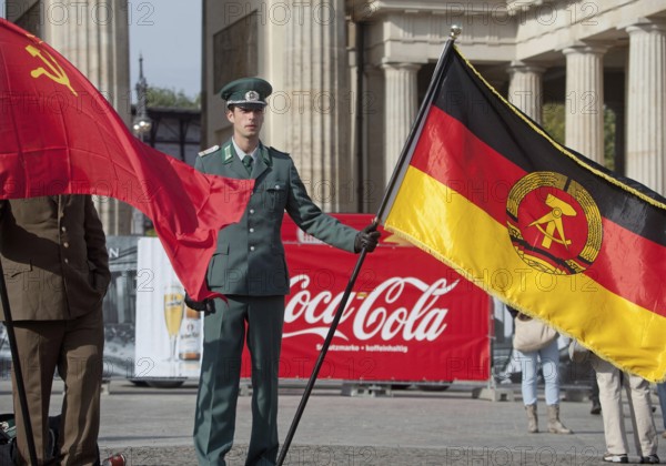 DEU Germany Berlin Actor wearing NVA uniform poses with the GDR flag in front of a Coca-Cola commercial. Scene on the eve of the 20th anniversary of German reunification on the festival mile in Berlin. Germany Berlin Actor in GDR Army Uniform Poses with GDR Flag in Front of a Coke Banner. Scene from the public party one day ahead of the celebration marking the 20th anniversary of the German reunification