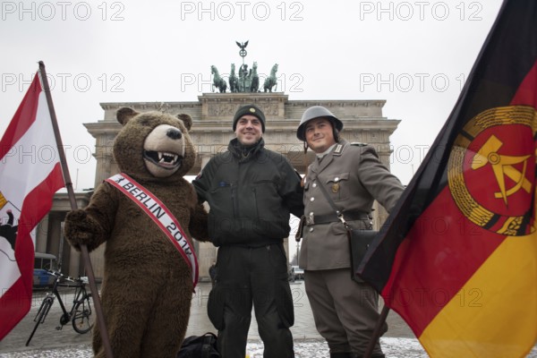 A riot police officer with the Berlin Bear and an actor in NVA uniform is photographed by a colleague in front of the Brandenburg Gate