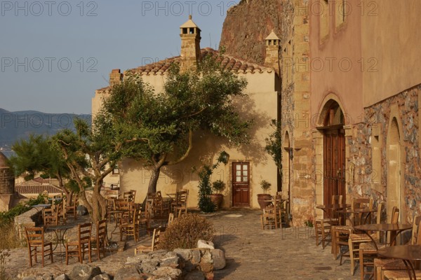 Soft morning light, a rustic outdoor café with tables and chairs on paved ground, sheltered by trees, Monemvasia, Monemvassia, Malvasy, Byzantine small town, rocky island, fortress, Laconia, Peloponnese, peninsula, Greece
