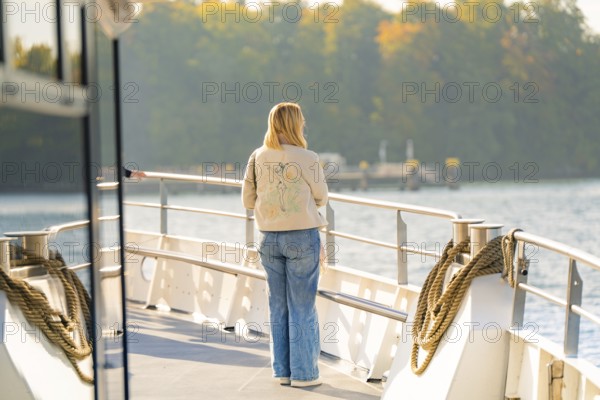 Woman on the deck of a boat looking at water and autumn trees in the distance, boat trip, Lake Constance, Germany
