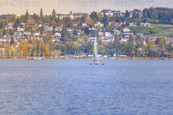 Sailing boat on a lake in front of a hilly landscape with colorful houses and autumn trees, boat trip, Lake Constance, Germany