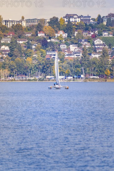 Closer view of a sailboat in front of a shore with colorful houses and autumn trees, boat trip, Lake Constance, Germany